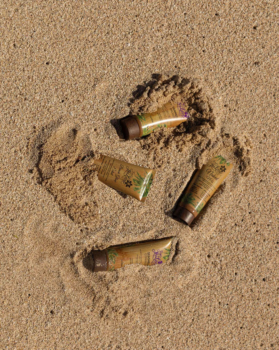 Four Le Lani CC cream tubes in various skin tones are partially buried in light brown, textured beach sand.
