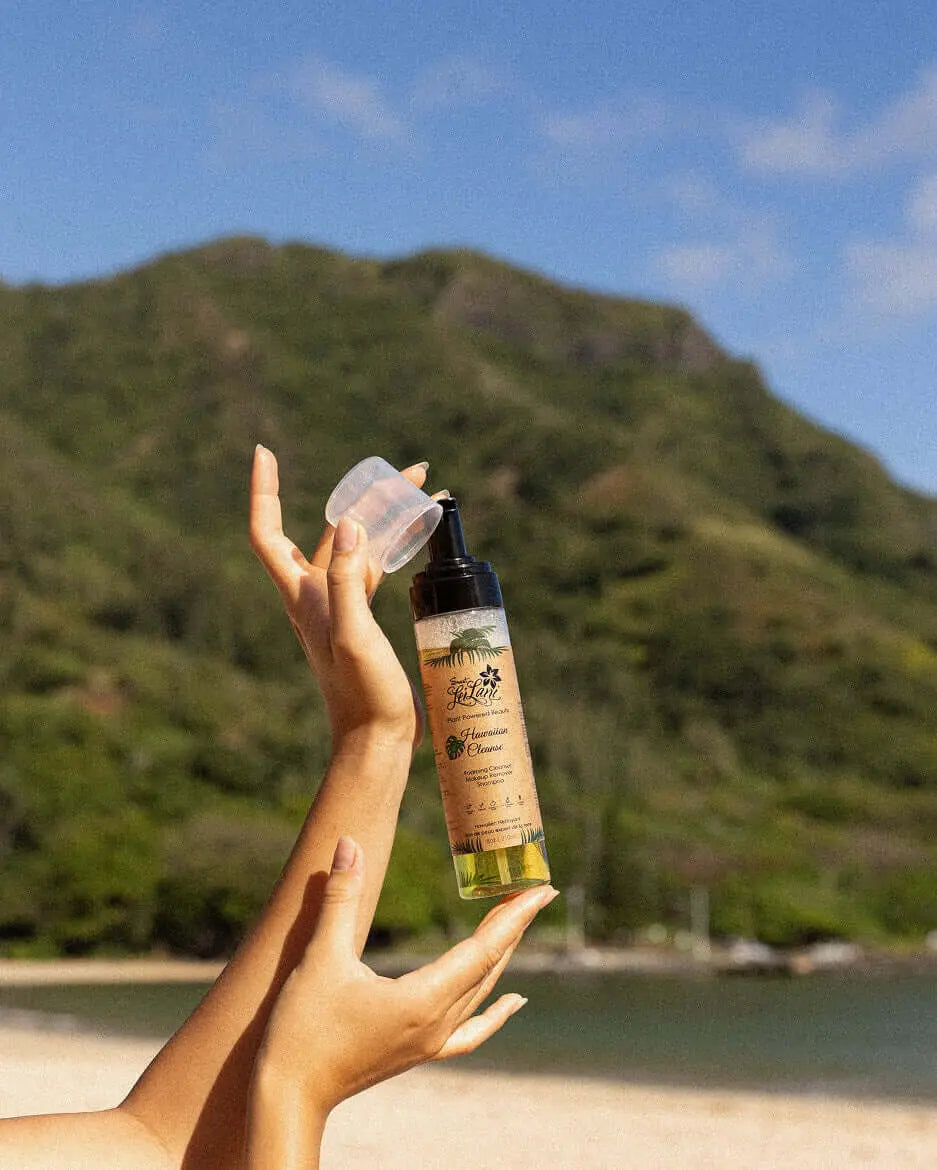 Hands holding a Sweet LeiLani Hawaiian Cleanser bottle with a clear cap, against a beach and mountain backdrop.