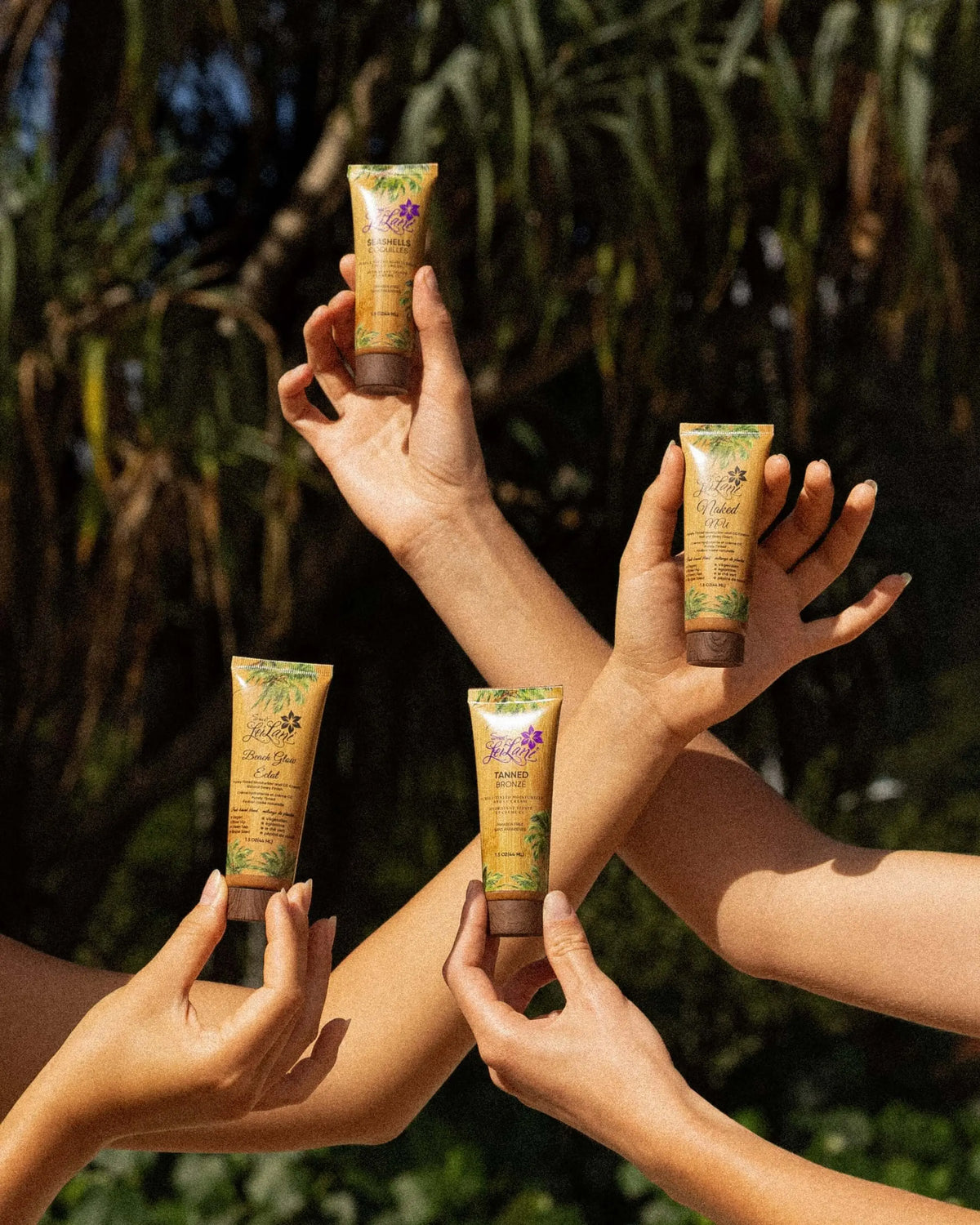 Hands holding four tubes of CC cream with tropical palm patterns, set against a blurred outdoor jungle background.