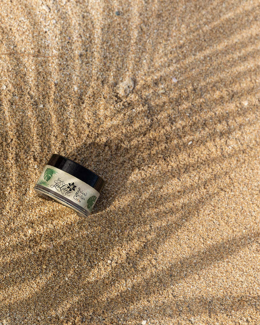Island Moisture face and hair conditioner jar resting on sandy beach with shadows.