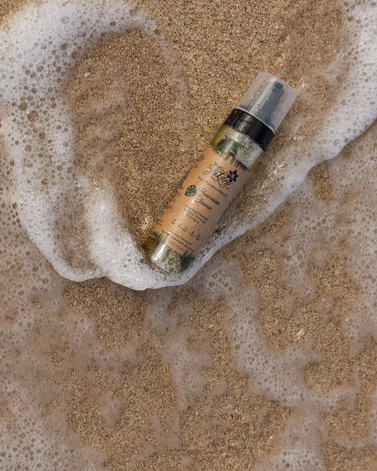 Sweet LeiLani Hawaiian Cleanser bottle with a pump lid resting on wet, foamy beach sand.