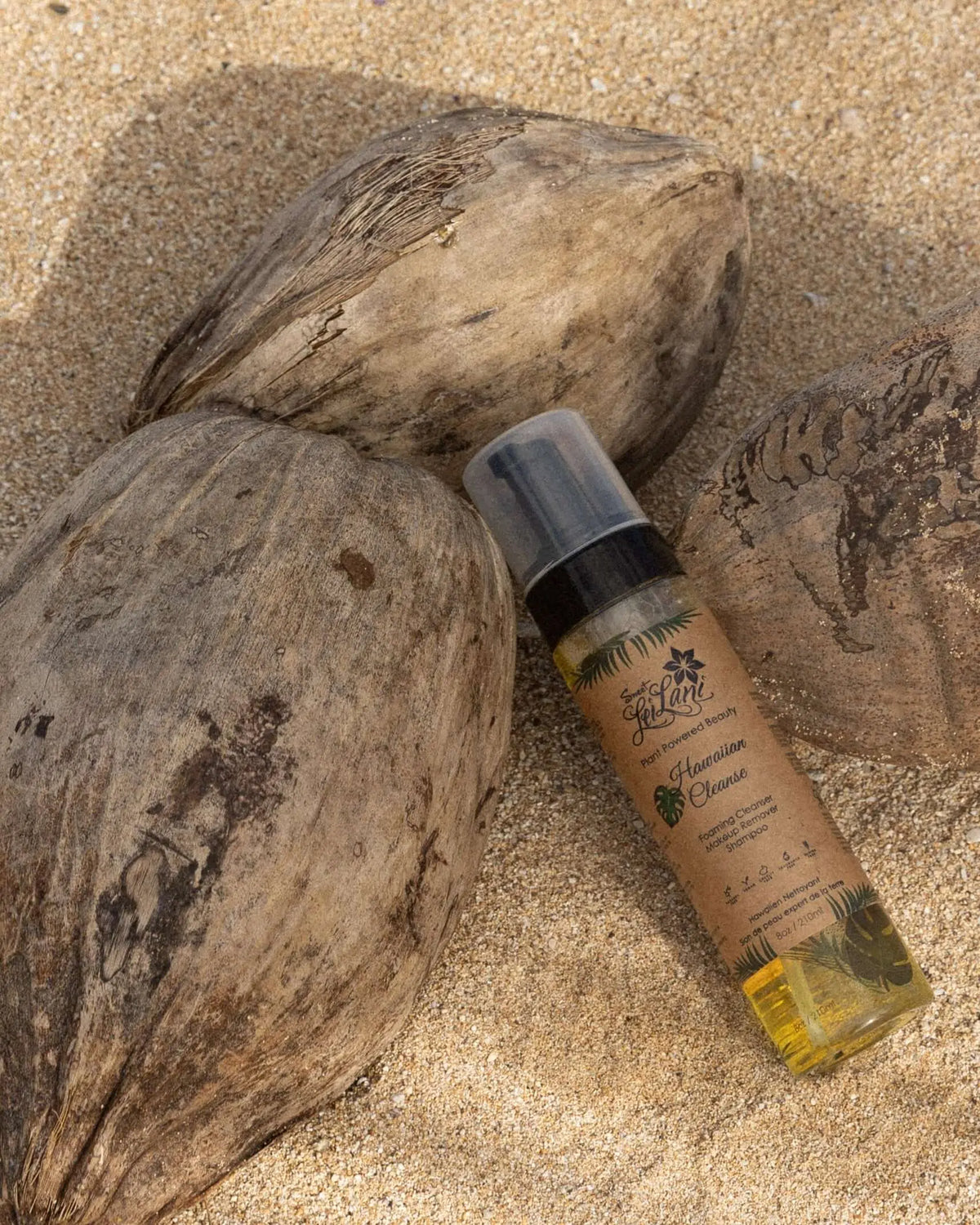 Sweet LeiLani Hawaiian Cleanse foaming pump bottle resting on sandy beach between textured brown coconuts.