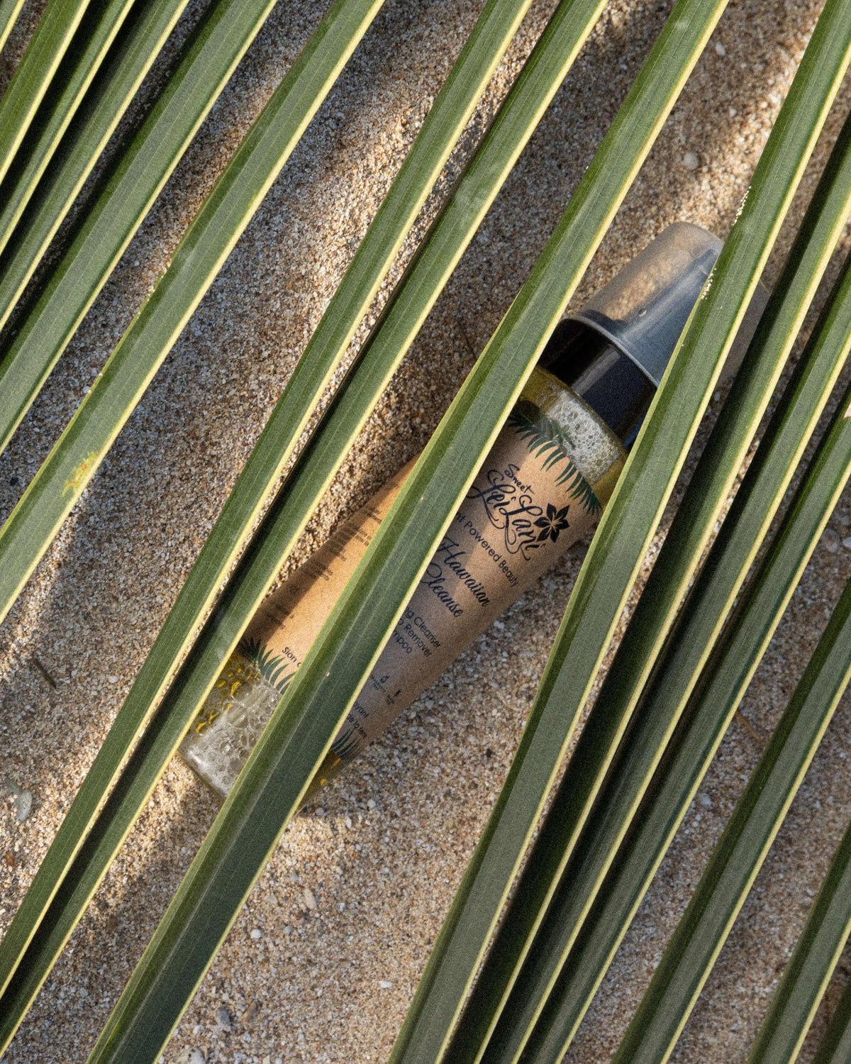 Sweet LeiLani Hawaiian Cleanser bottle placed on sand and partially obscured by green palm fronds.
