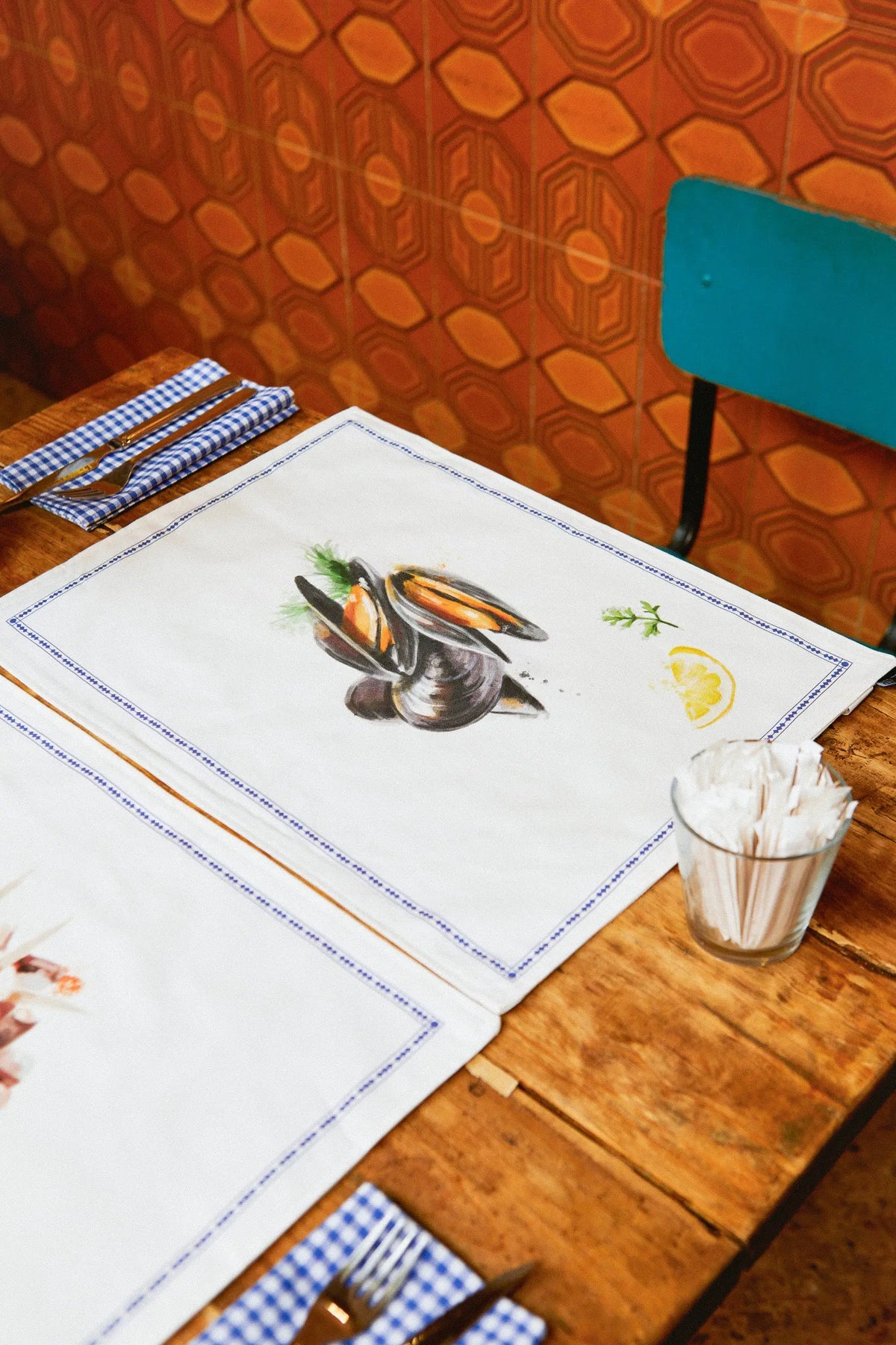 White reversible placemats featuring a watercolor mussel and lemon illustration, blue border, on a wooden table.
