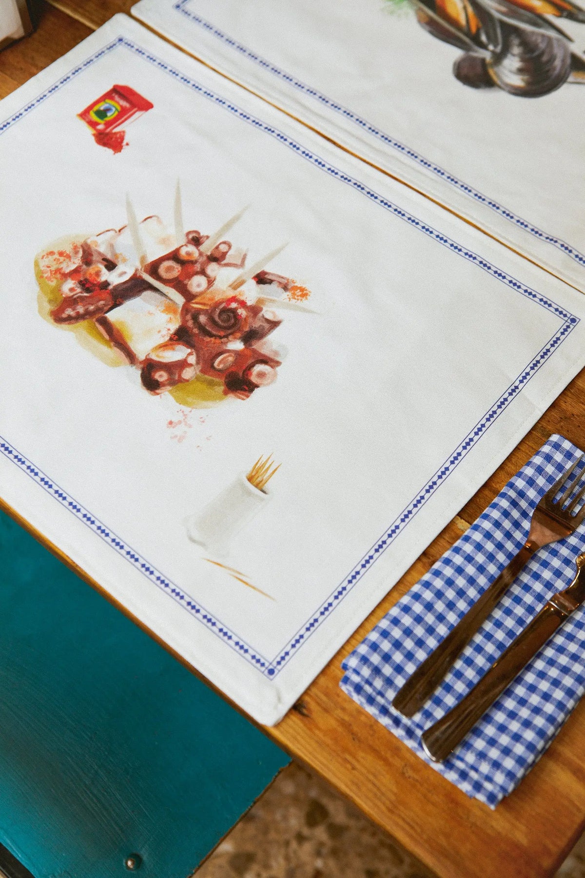 Dos manteles individuales de tela con estampado de pulpo, marco azul y servilleta de cuadros sobre mesa de madera.