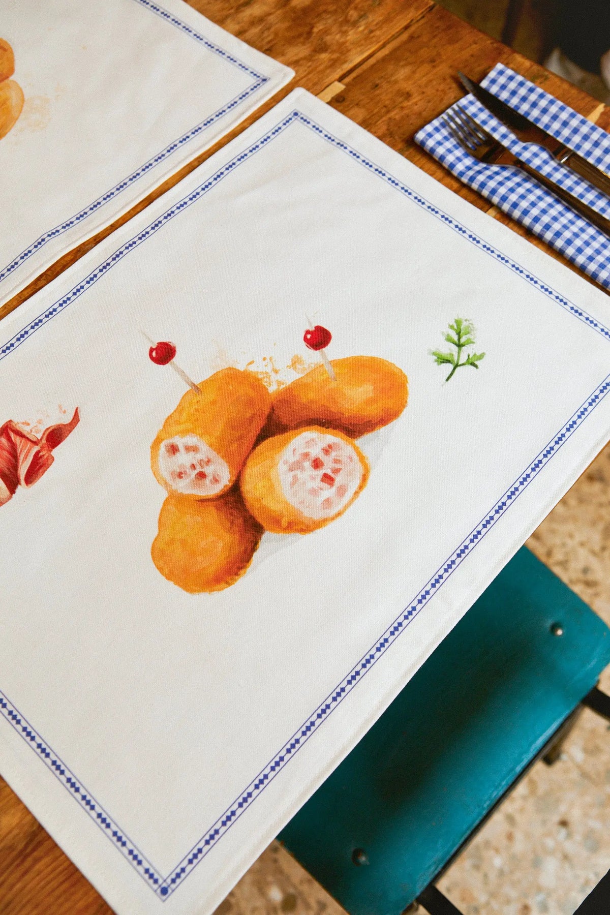 Pack de dos manteles individuales blancos con estampado de croquetas y borde azul, sobre una mesa de madera.