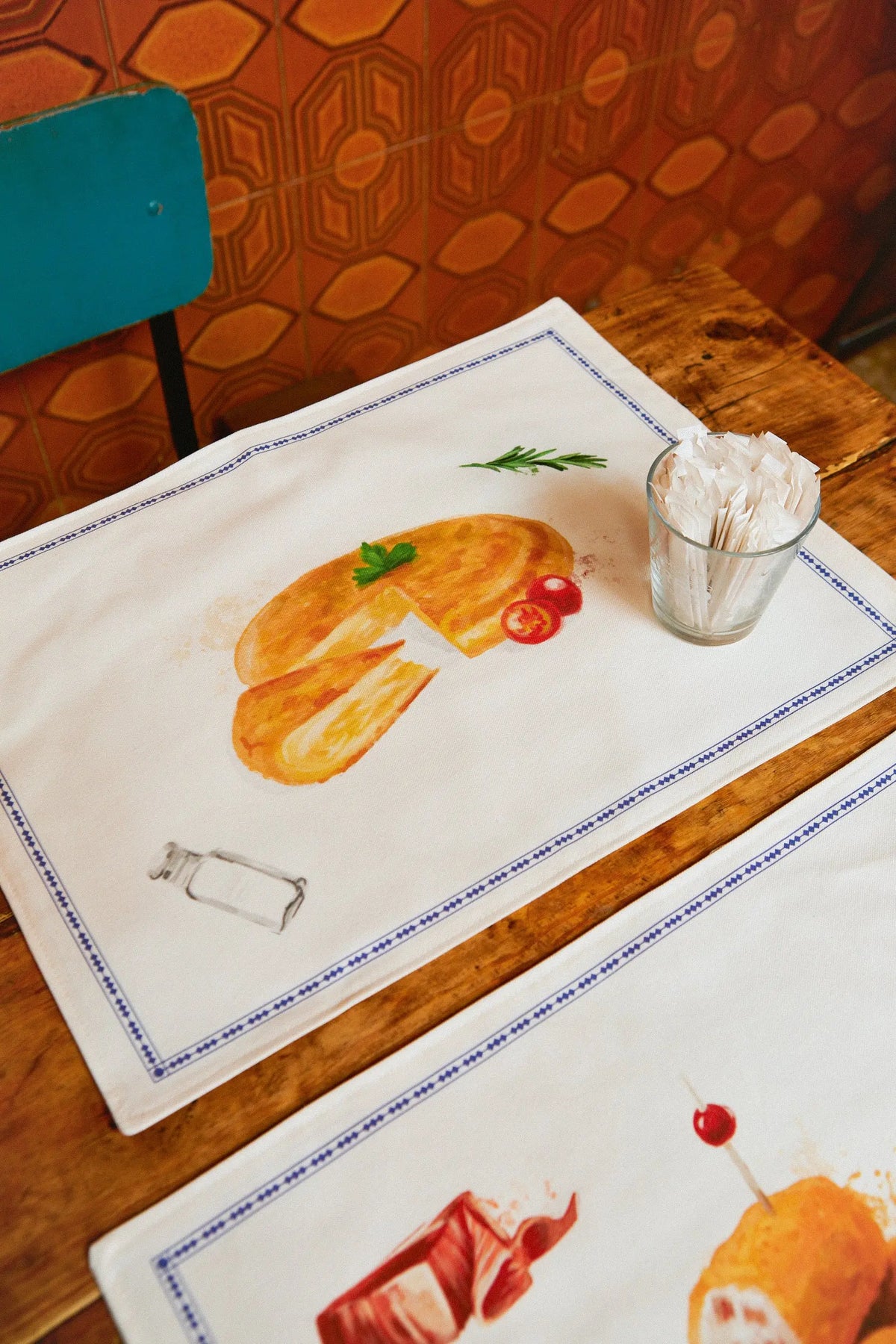 Pack de 2 manteles individuales de tela con estampado de tapas españolas y borde azul sobre mesa de madera.