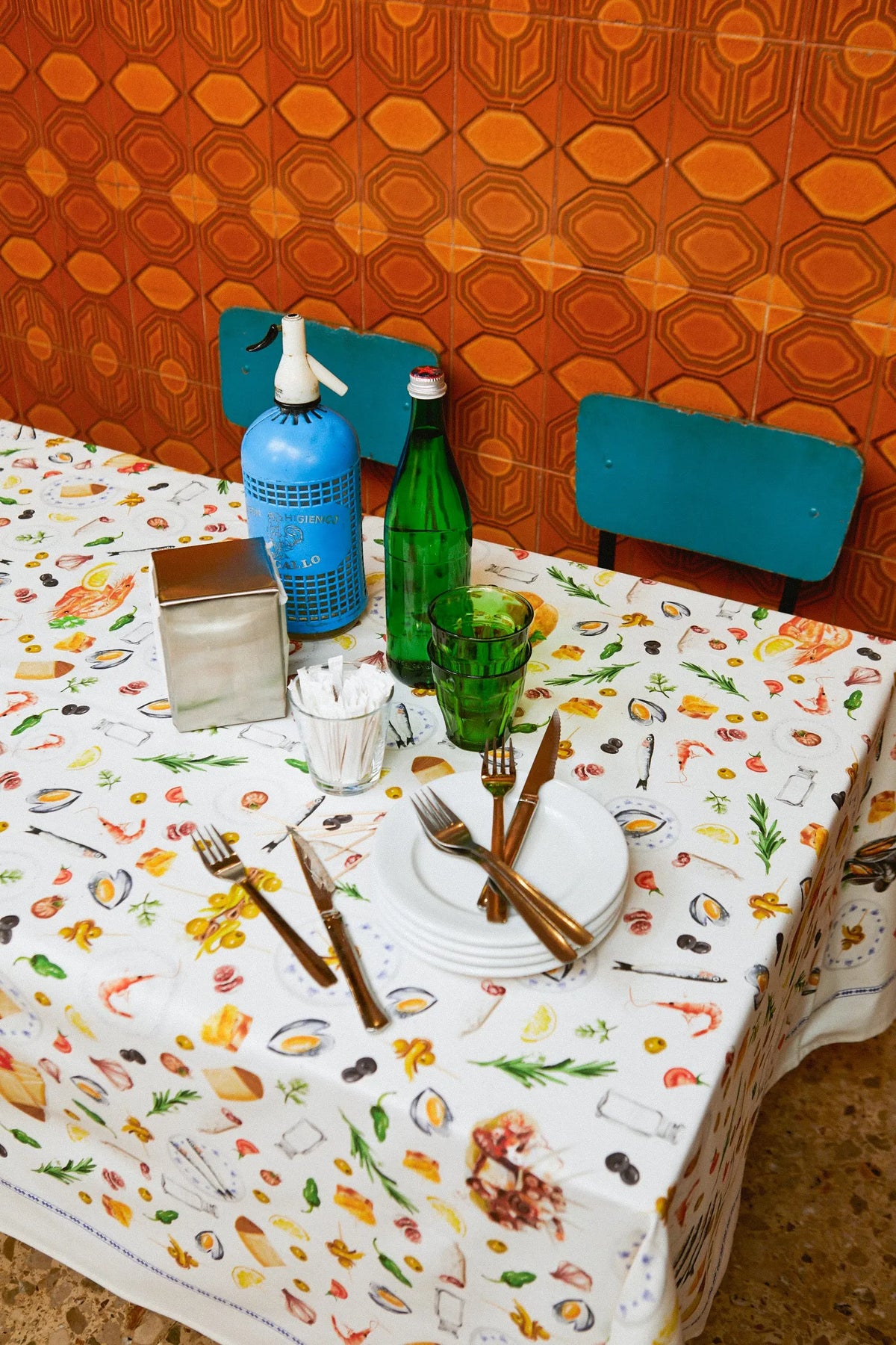 Mantel blanco con estampado de tapas, mariscos y cubiertos. Mesa puesta con vajilla, cristalería y sifón azul.