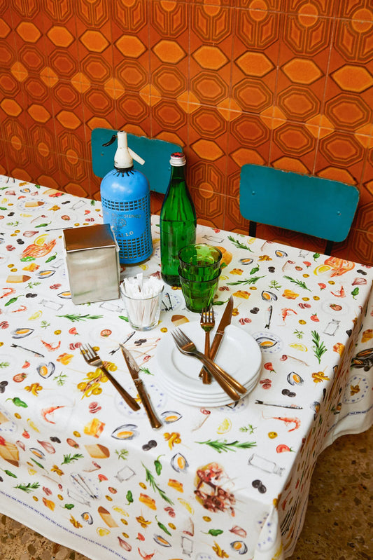 Mantel blanco con estampado de tapas, mariscos y cubiertos. Mesa puesta con vajilla, cristalería y sifón azul.