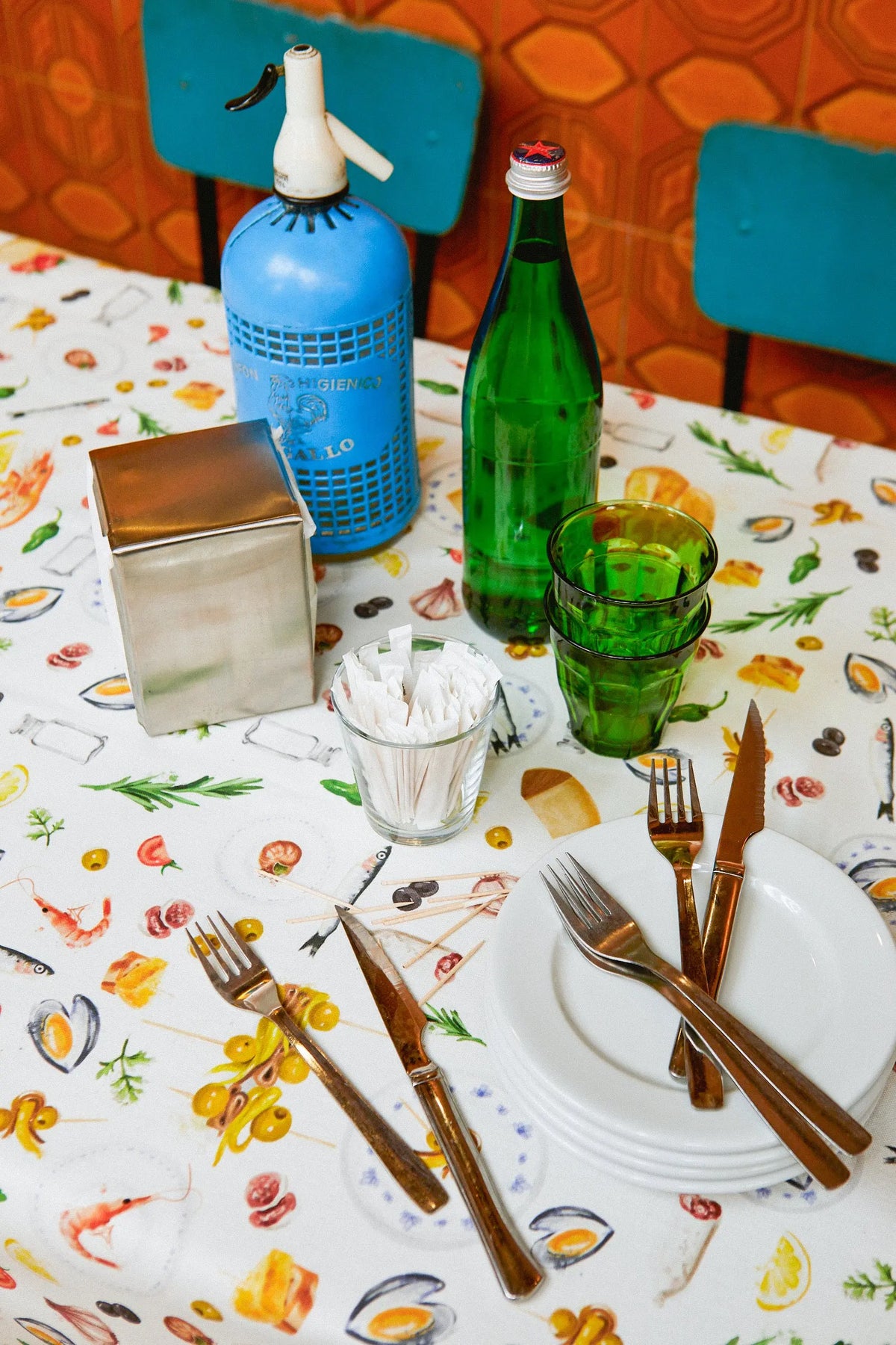 Mantel blanco con estampado de tapas, cubiertos metálicos, vajilla y cristalería sobre una mesa de bar.