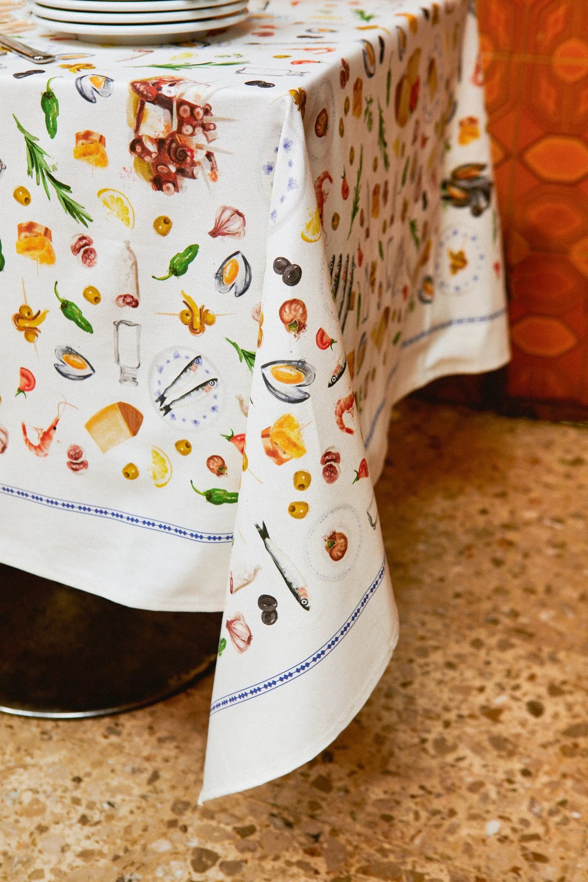 Mantel blanco con estampado de tapas españolas, ingredientes y mariscos, con cenefa azul geométrica en el borde.