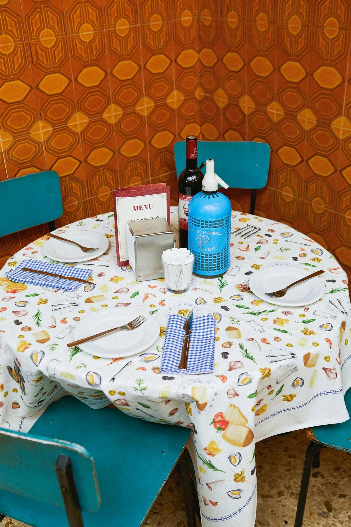 Mantel blanco redondo con estampado colorido de tapas y alimentos, servilletas de cuadros azules y vajilla básica.