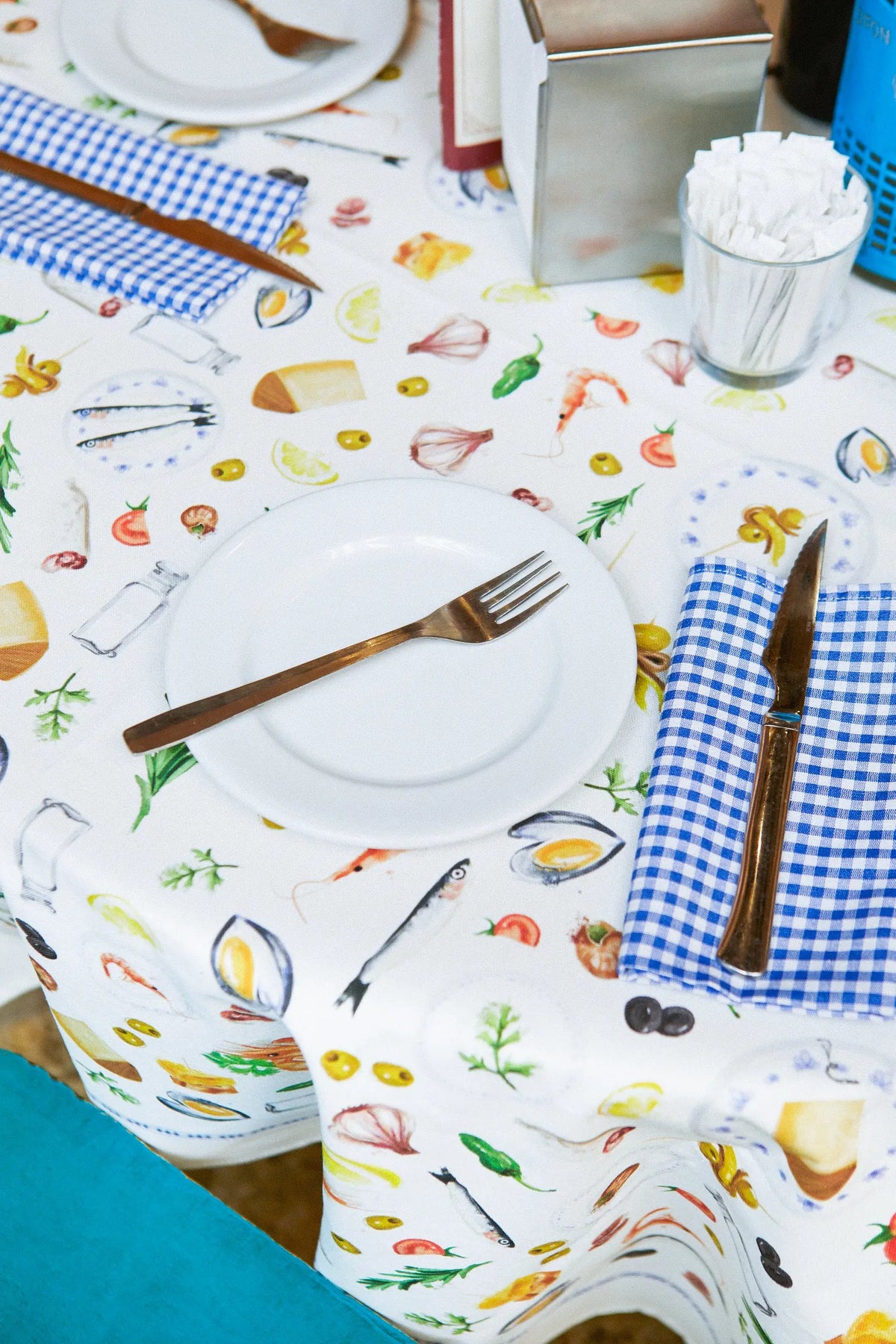 Mantel blanco con estampado de tapas e ingredientes mediterráneos. Incluye servilletas de cuadros azules y vajilla.