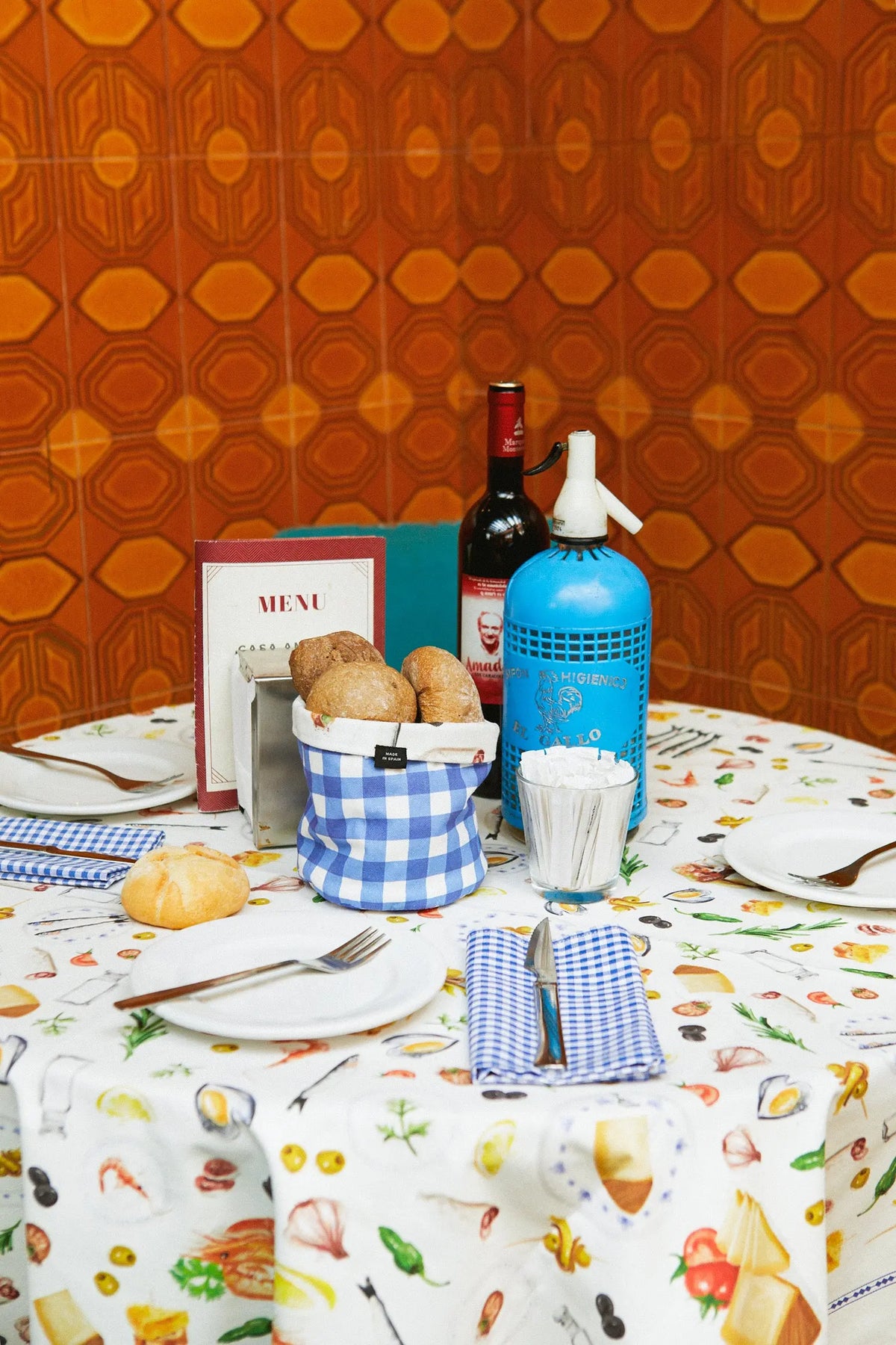 Mantel blanco estampado con ilustraciones de tapas y comida, colocado en una mesa con vajilla y servilletas de cuadros.
