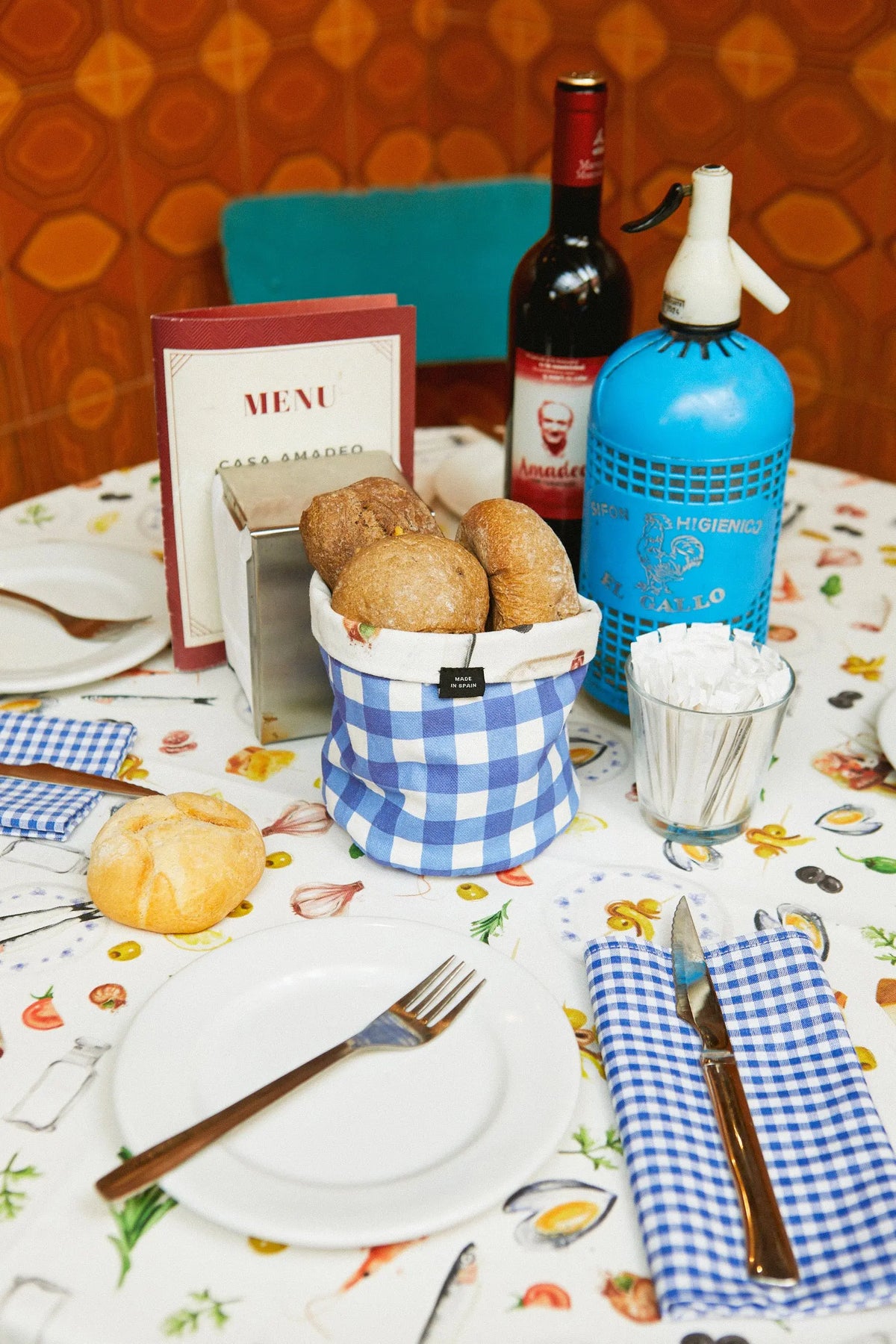 Mantel blanco con estampado de alimentos y servilletas de tela a cuadros azules sobre mesa con pan y cubiertos.