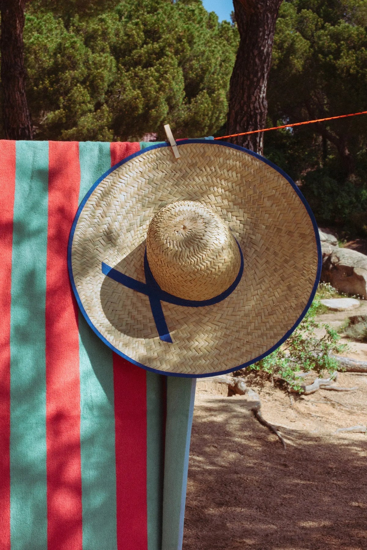 Sombrero de paja con cinta azul, colgado sobre toalla de playa rayada en rojo y verde, estilo veraniego.