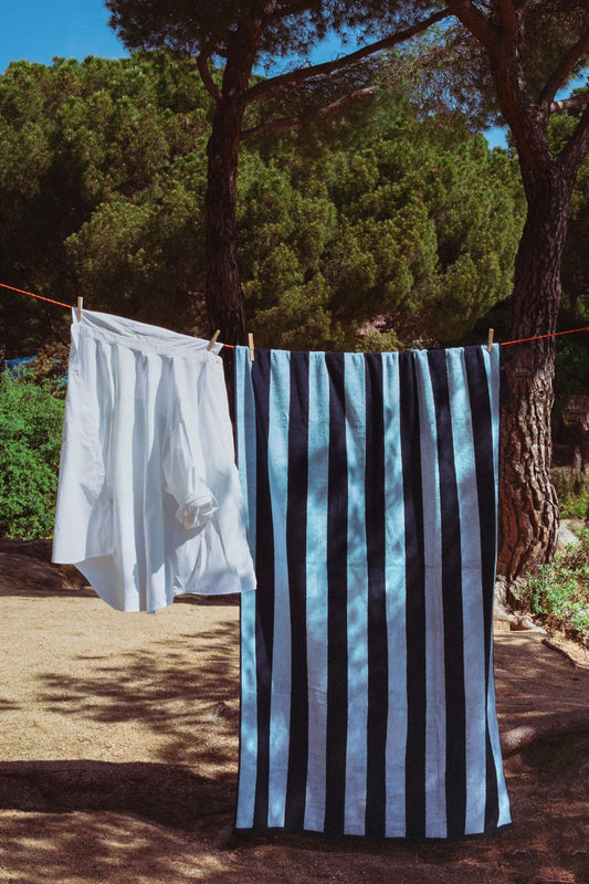 Toalla de playa grande con rayas verticales en azul marino y celeste, colgada de un tendedero al aire libre.