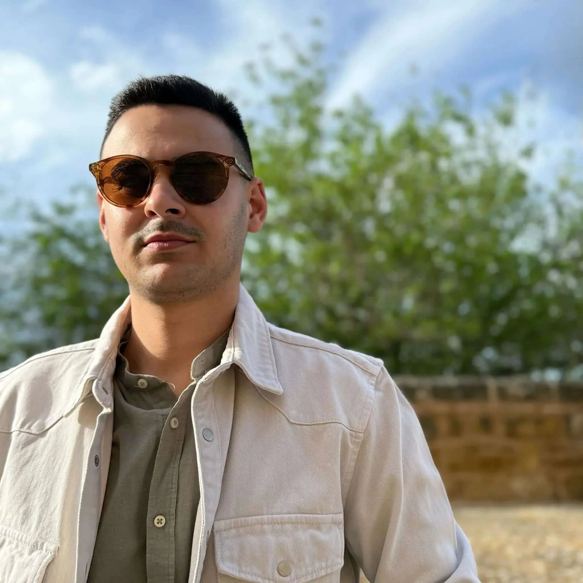 Hombre con gafas de sol redondas de montura naranja cristal y lentes marrones en exteriores con chaqueta beige.