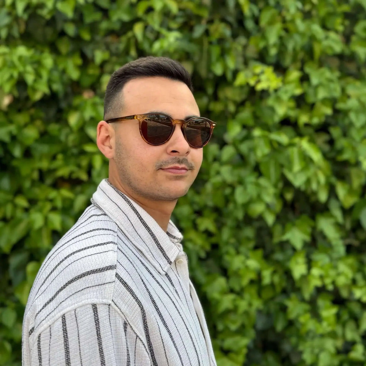 Hombre con gafas de sol redondas de acetato naranja transparente y lentes marrones, con camisa de rayas, ante hiedra.