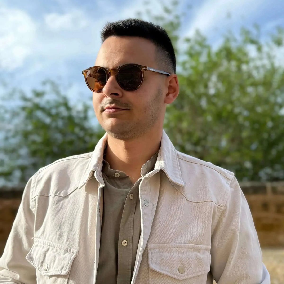 Hombre con gafas de sol redondas de montura naranja cristal y lentes marrones, vistiendo camisa y sobrecamisa beis.