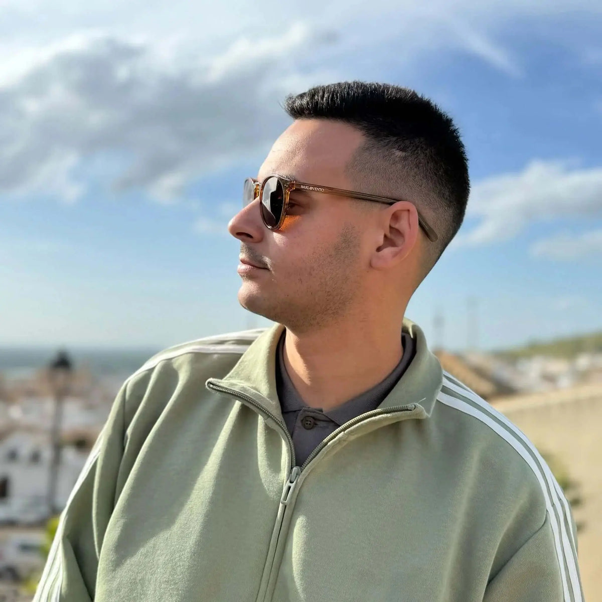 Joven con gafas de sol redondas de montura naranja cristal y lentes marrones, vistiendo chaqueta deportiva verde oliva.