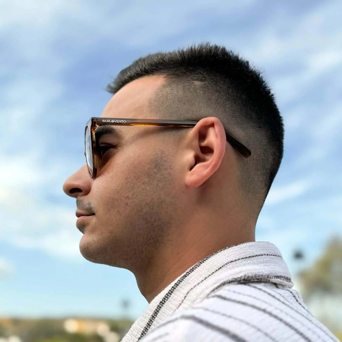 Hombre de perfil con gafas de sol redondas de montura naranja translúcida y lentes marrones sobre fondo de cielo.
