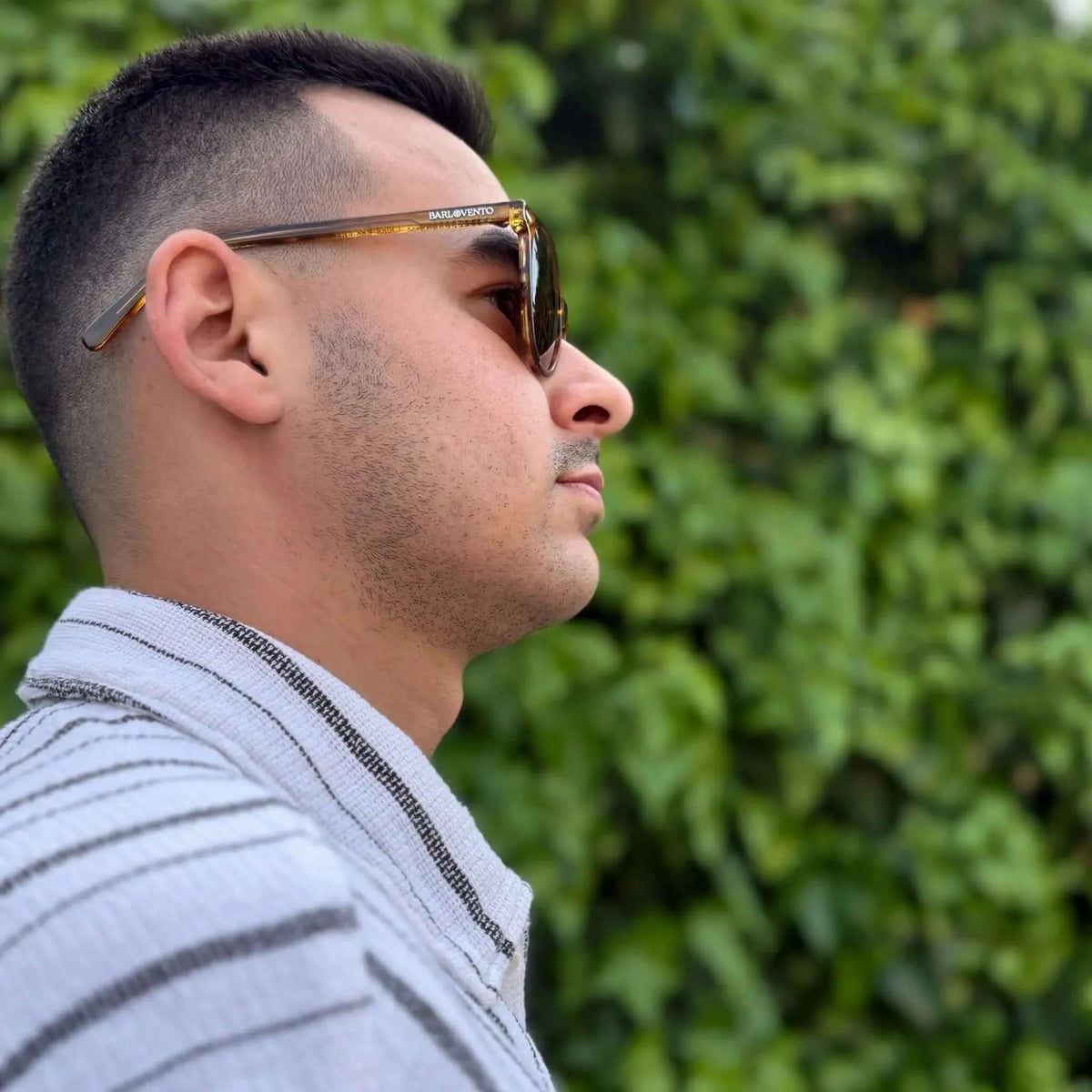 Hombre de perfil con gafas de sol redondas de acetato naranja translúcido y lentes marrones ante un fondo de hojas.