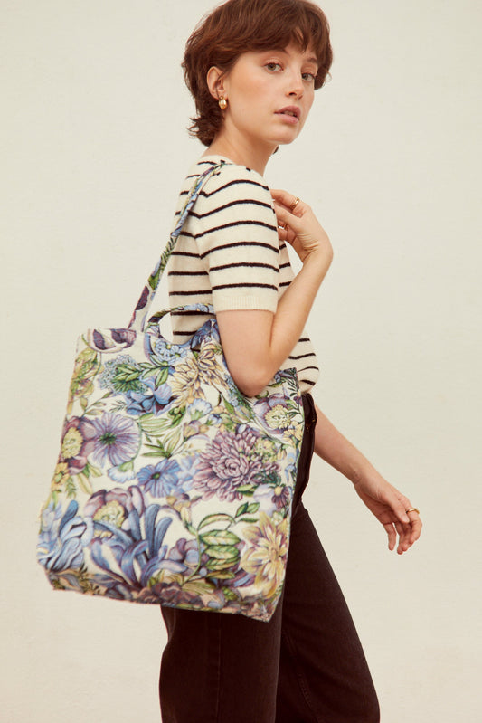 Tote bag de tela jacquard con estampado de crisantemos azules y tonos vegetales, llevada al hombro por una mujer.