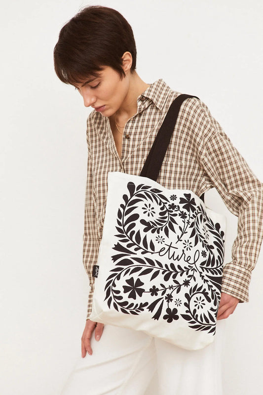 Tote bag blanco de tela con estampado floral y orgánico en color negro. Tiene asas negras y diseño cuadrado.