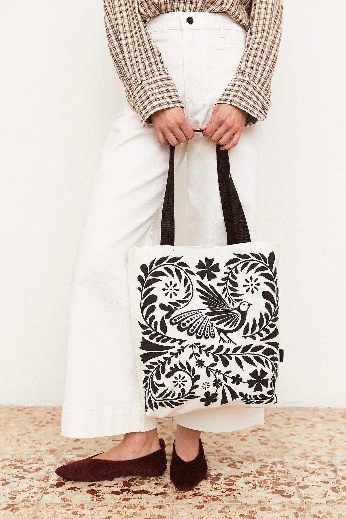 Tote bag de tela blanca con estampado de estilo folclórico en color negro, con un ave rodeada de flores y hojas.