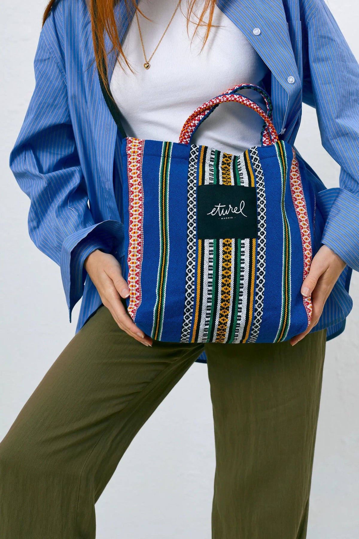 Bolso bandolera tipo tote de tela azul con estampado de rayas verticales étnicas multicolores y asas cortas tejidas.