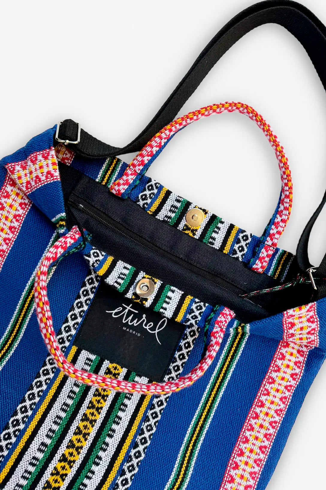 Bolso bandolera de tela azul con estampado étnico, asas tejidas multicolores, cierre de cremallera y botón a presión.