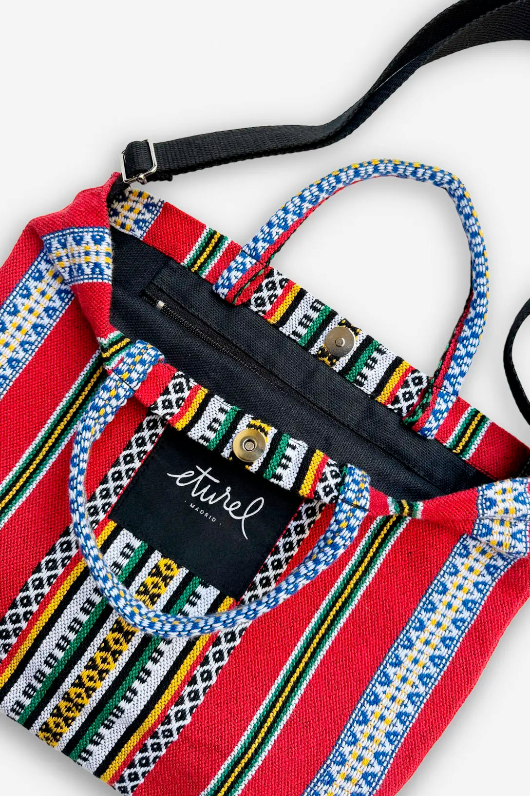 Bolso bandolera de tela roja con rayas multicolores, asas de mano, correa negra y cierre de botón a presión.