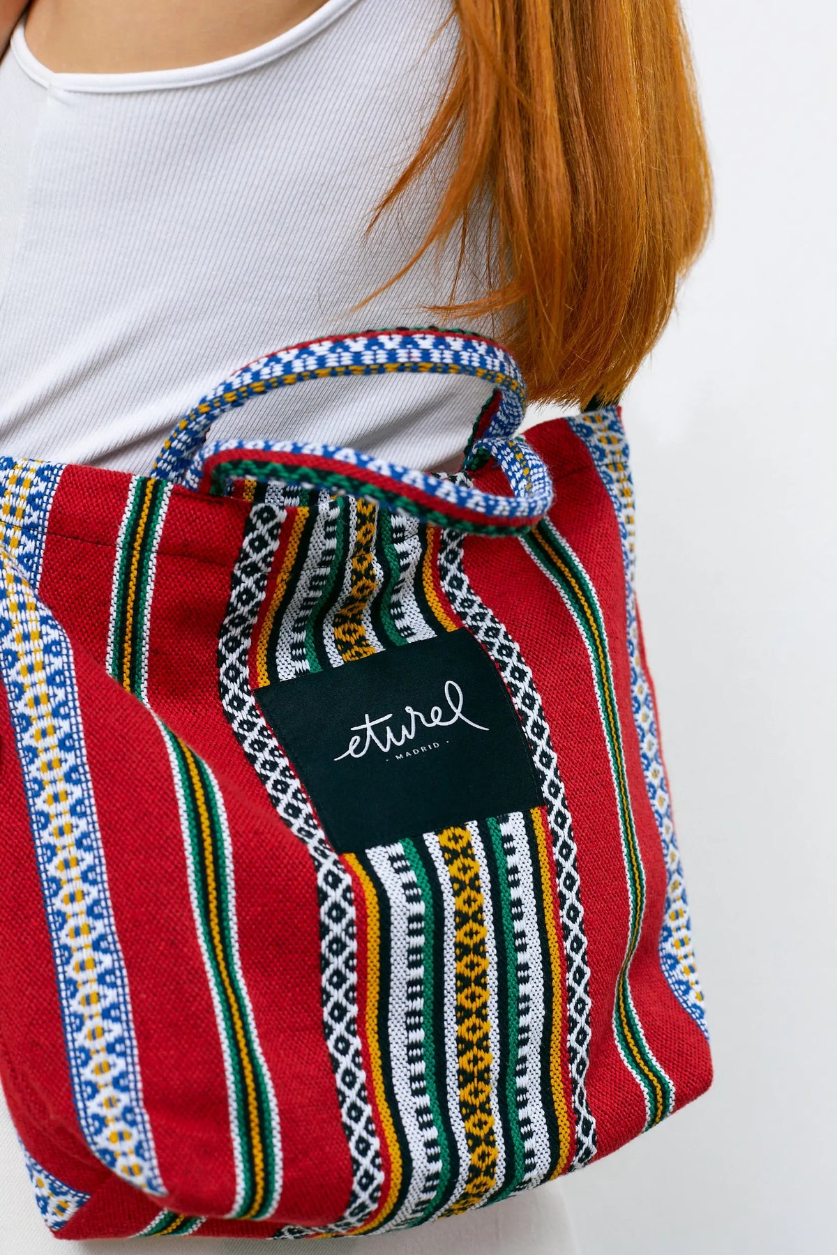 Bolso bandolera de tela roja con rayas multicolores, tejido texturizado y asa ajustable sobre el hombro.