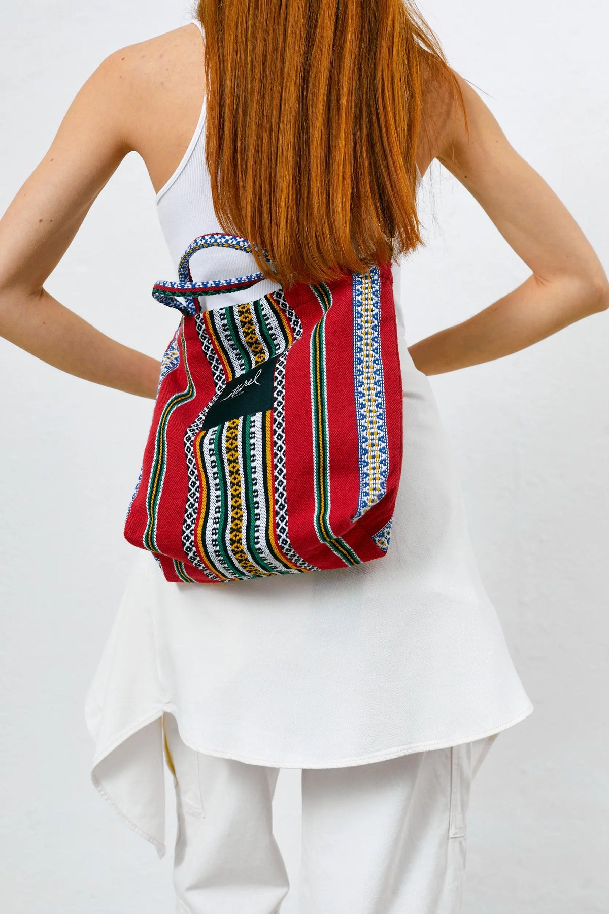 Bolso bandolera rojo de tela con estampado de rayas verticales multicolores y asas cortas, llevado en la espalda.