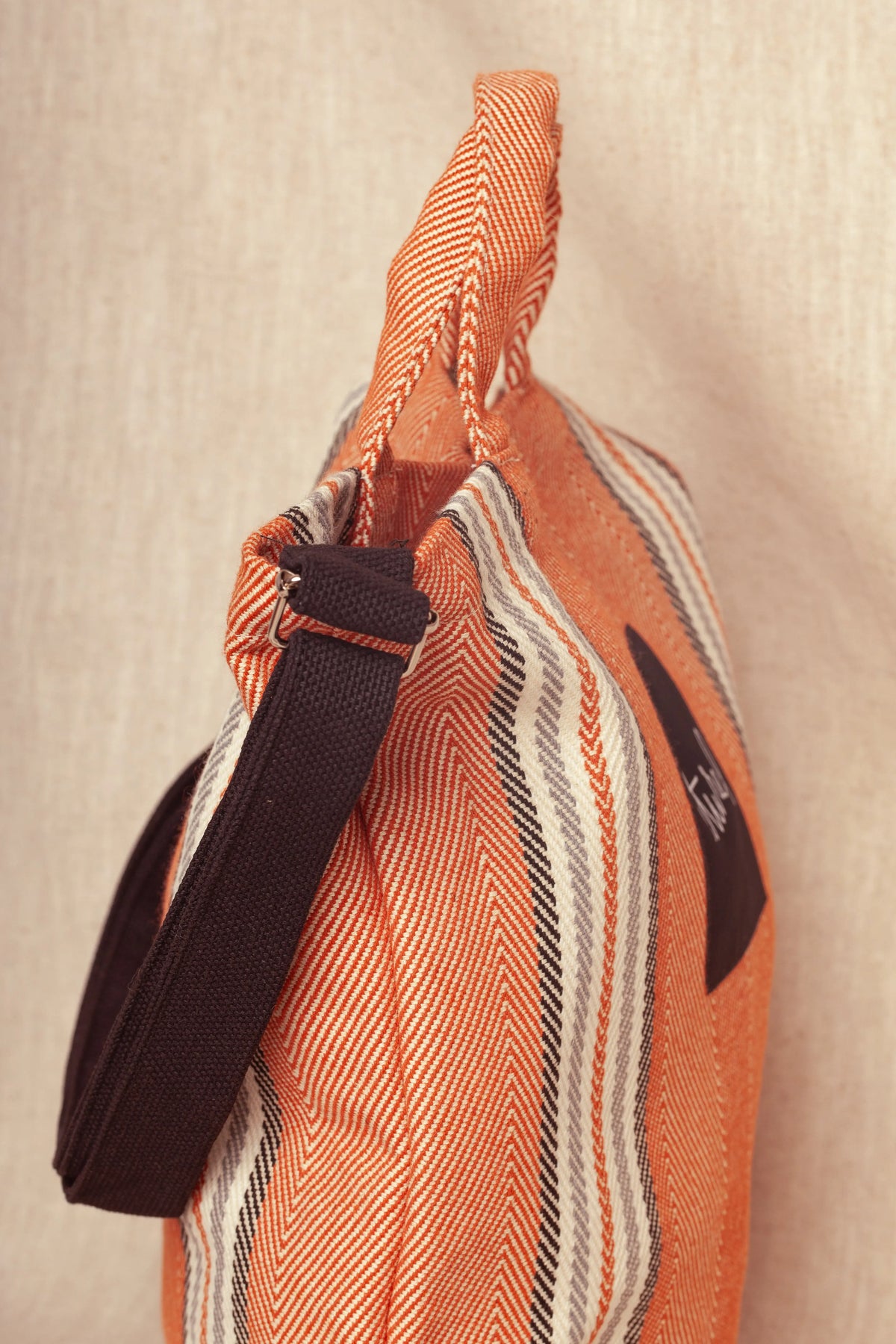 Bolso bandolera de sarga color arcilla con patrón de rayas y correa ajustable negra sobre fondo de tela clara.