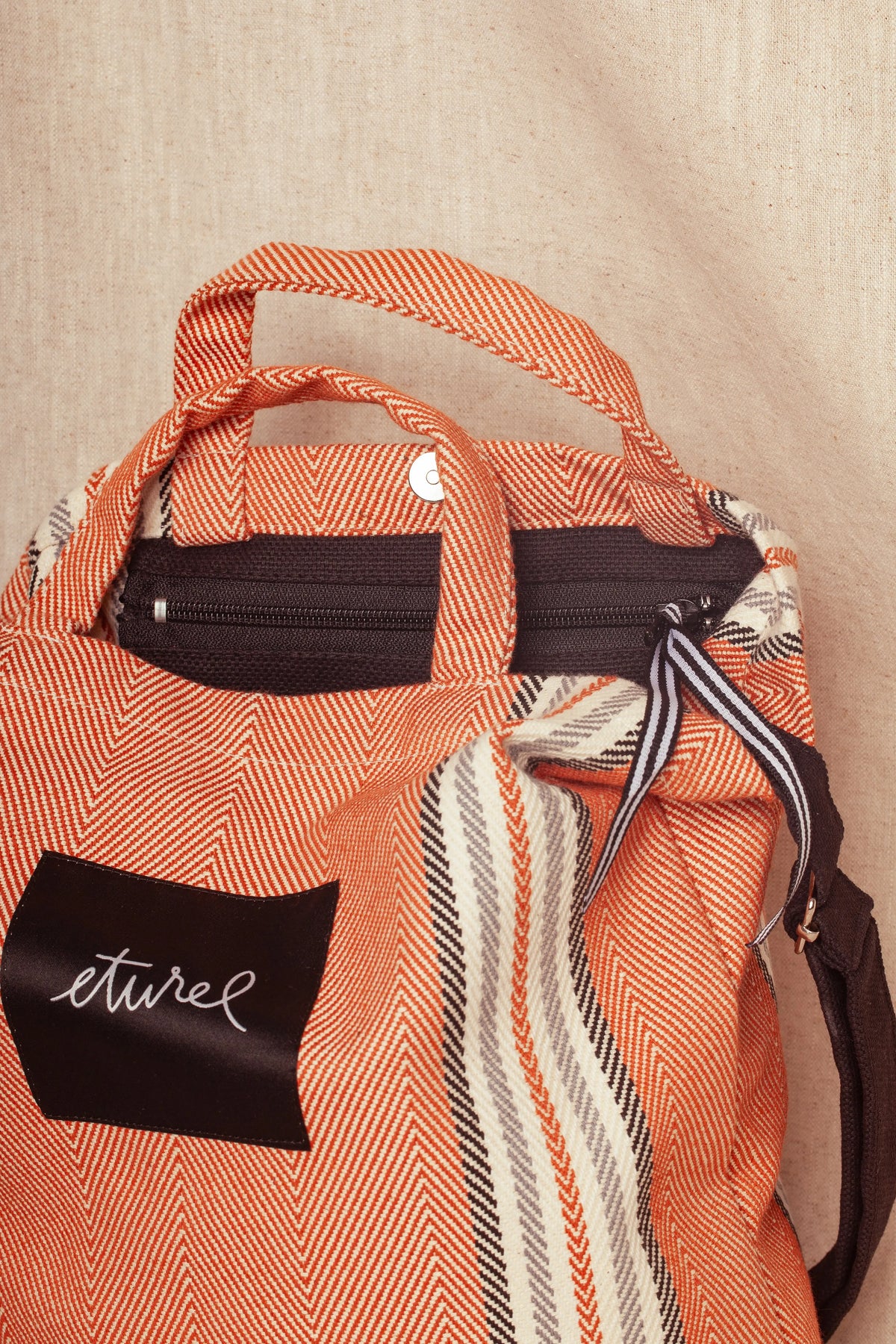 Clay-colored herringbone twill crossbody bag with dual handles, black zip closure, and branded black fabric label.