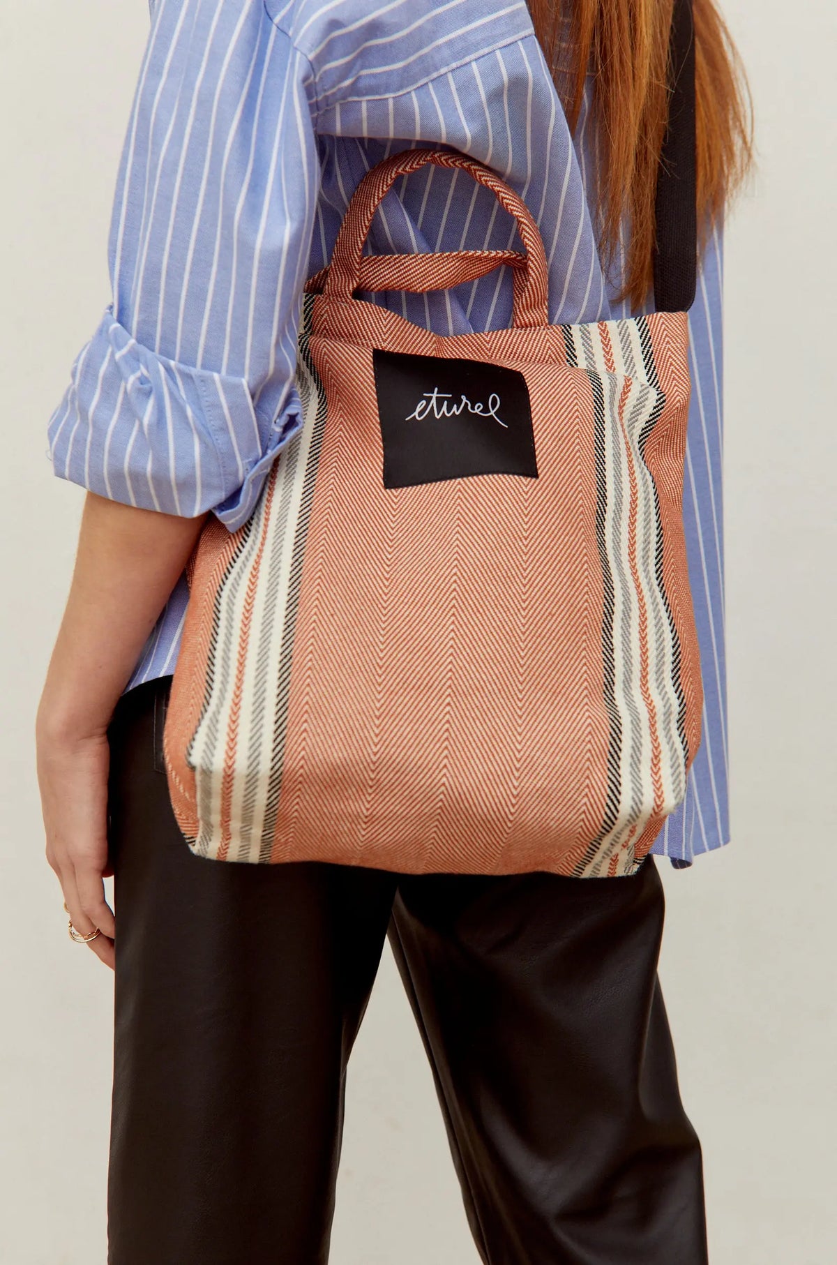 Clay-colored herringbone twill crossbody tote bag with black and white side stripes and top handles.