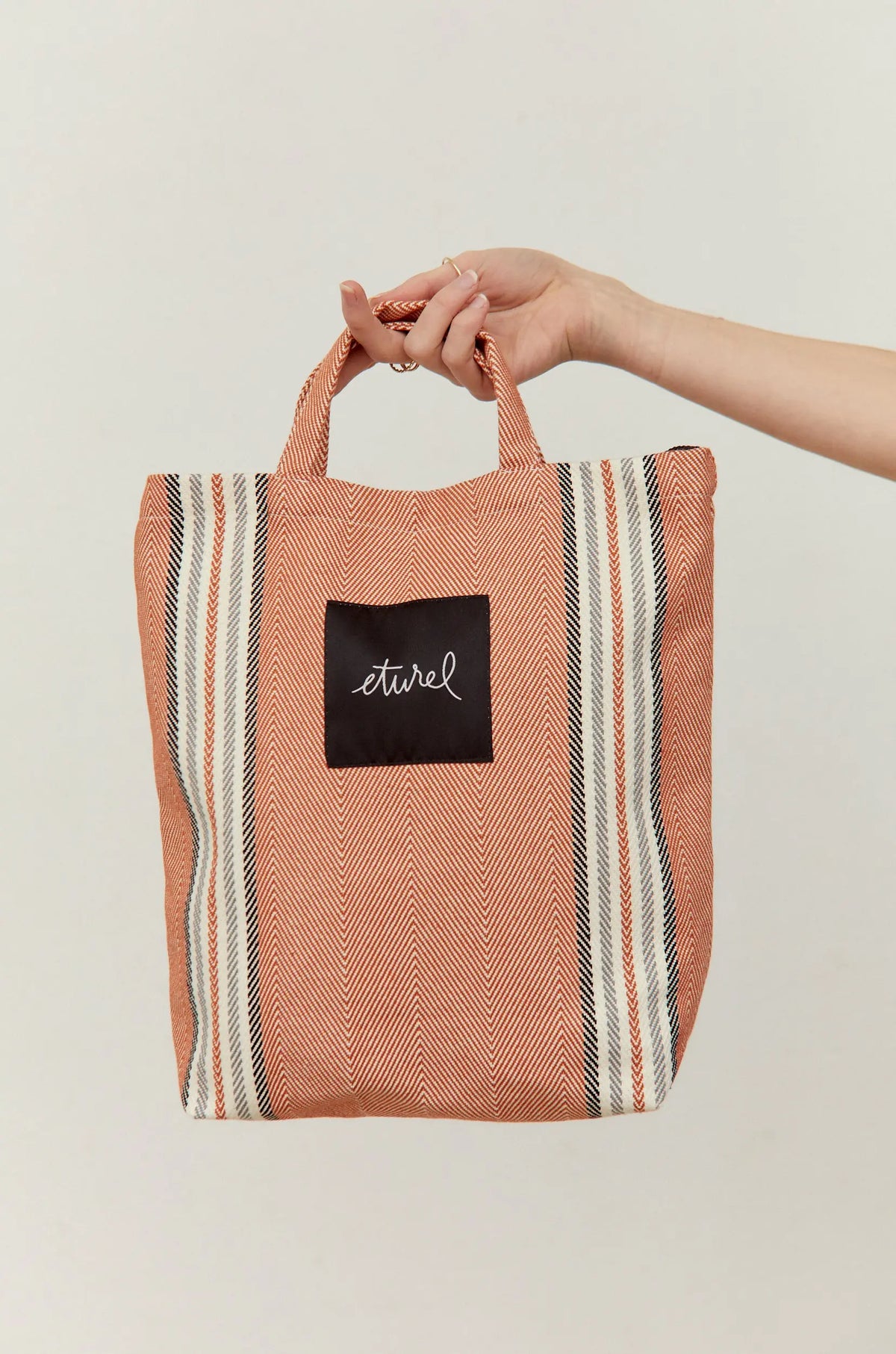 Tote bag bandolera de sarga color arcilla con estampado de rayas verticales y tejido en espiga, sostenida por una mano.