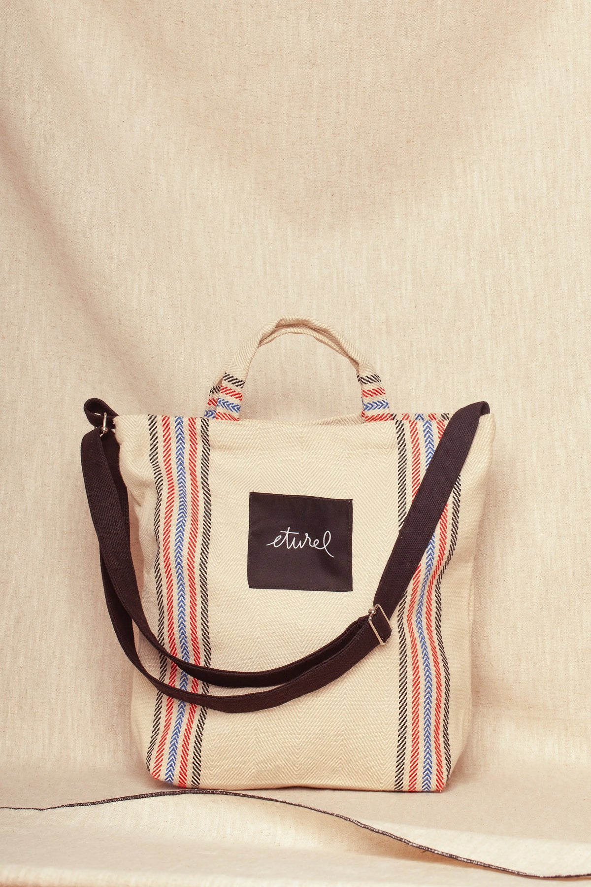 Bolso bandolera de sarga color crudo con rayas verticales rojas y azules y correa ajustable negra sobre fondo neutro.