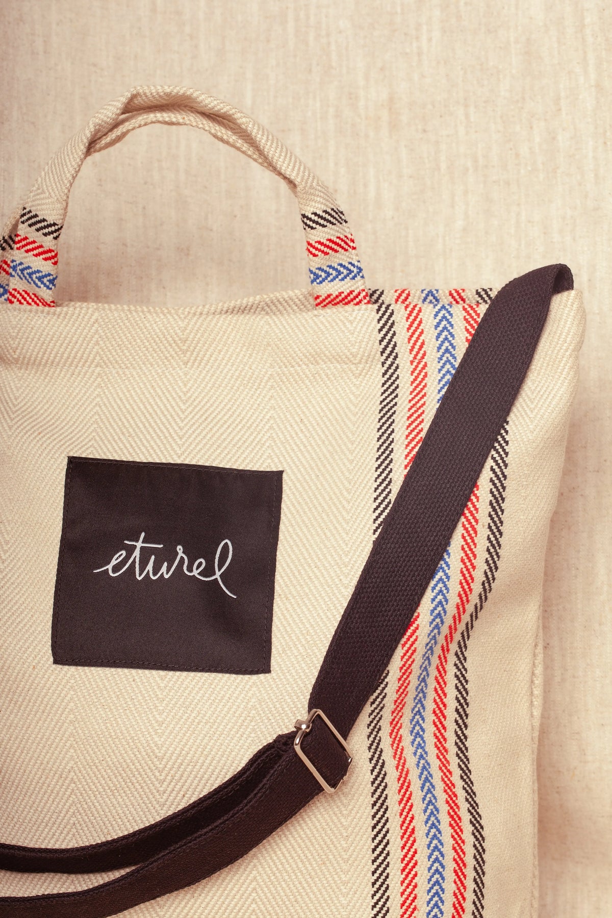 Bolso bandolera de sarga color crudo con textura de espiga, rayas rojas y azules, asas cortas y correa negra ajustable.