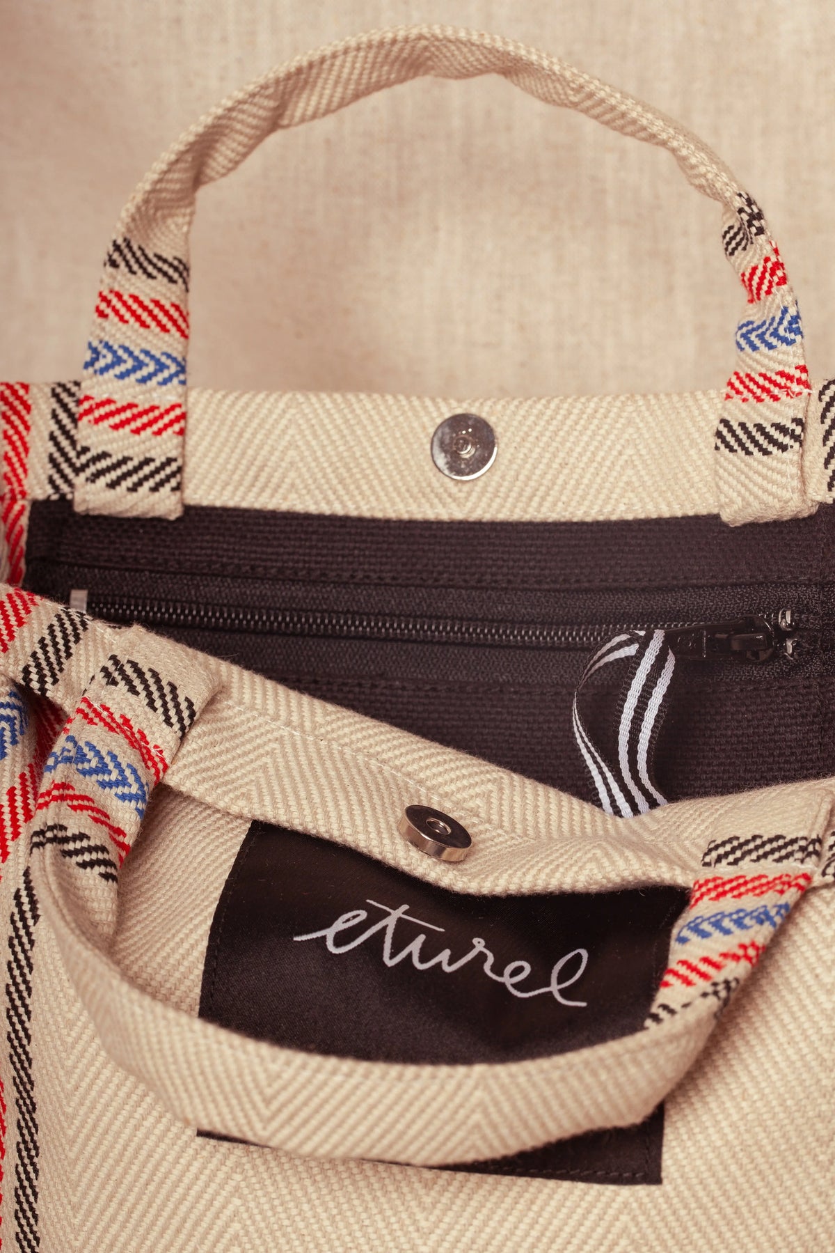 Cream herringbone twill tote with red, blue, and black striped handles, magnetic clasp, and interior zip pocket.