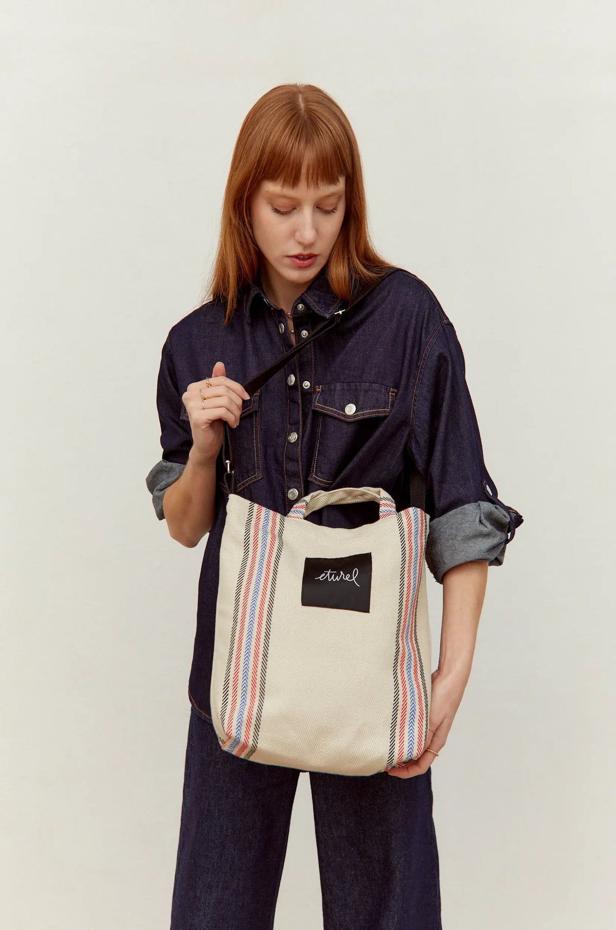 Bolso bandolera tipo tote de sarga cruda con rayas laterales rojas y azules, llevado al hombro sobre camisa denim.