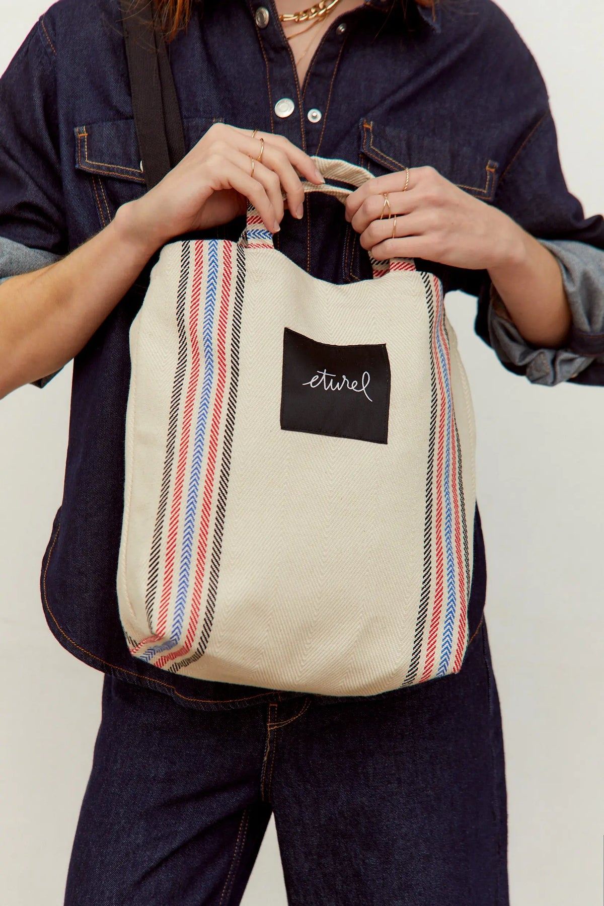Bolso bandolera tipo tote en sarga cruda con estampado de rayas verticales rojas, azules y negras y asas cortas.