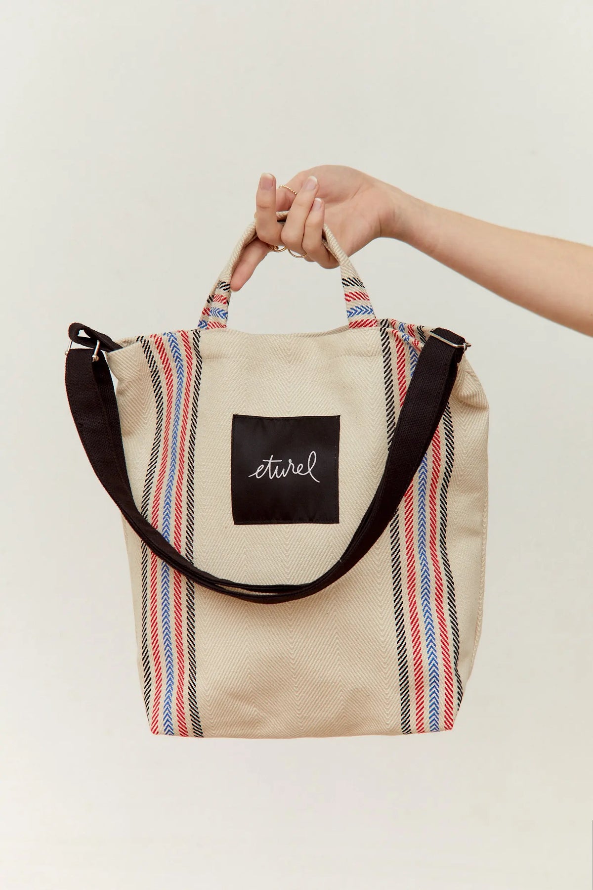Bolso bandolera tipo tote de sarga cruda con rayas rojas, azules y negras, asas cortas y correa negra ajustable.
