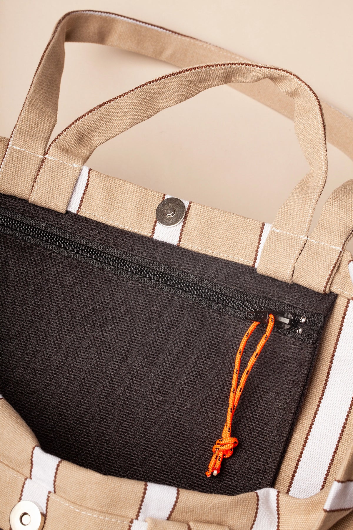 Bolso tote de lona a rayas beige con asa, cierre de botón magnético y bolsillo interior negro con cremallera naranja.