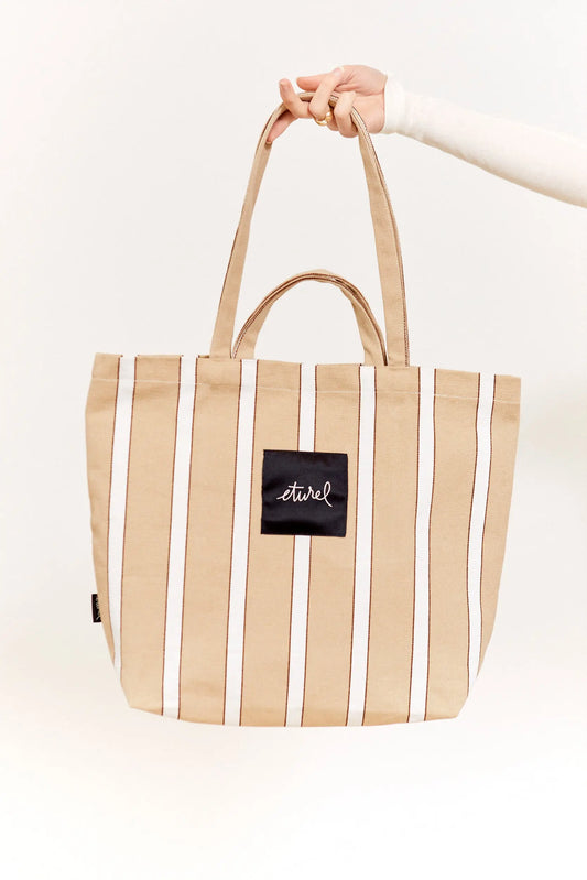 Beige striped canvas tote bag with dual handles and a central black logo patch held by a hand.