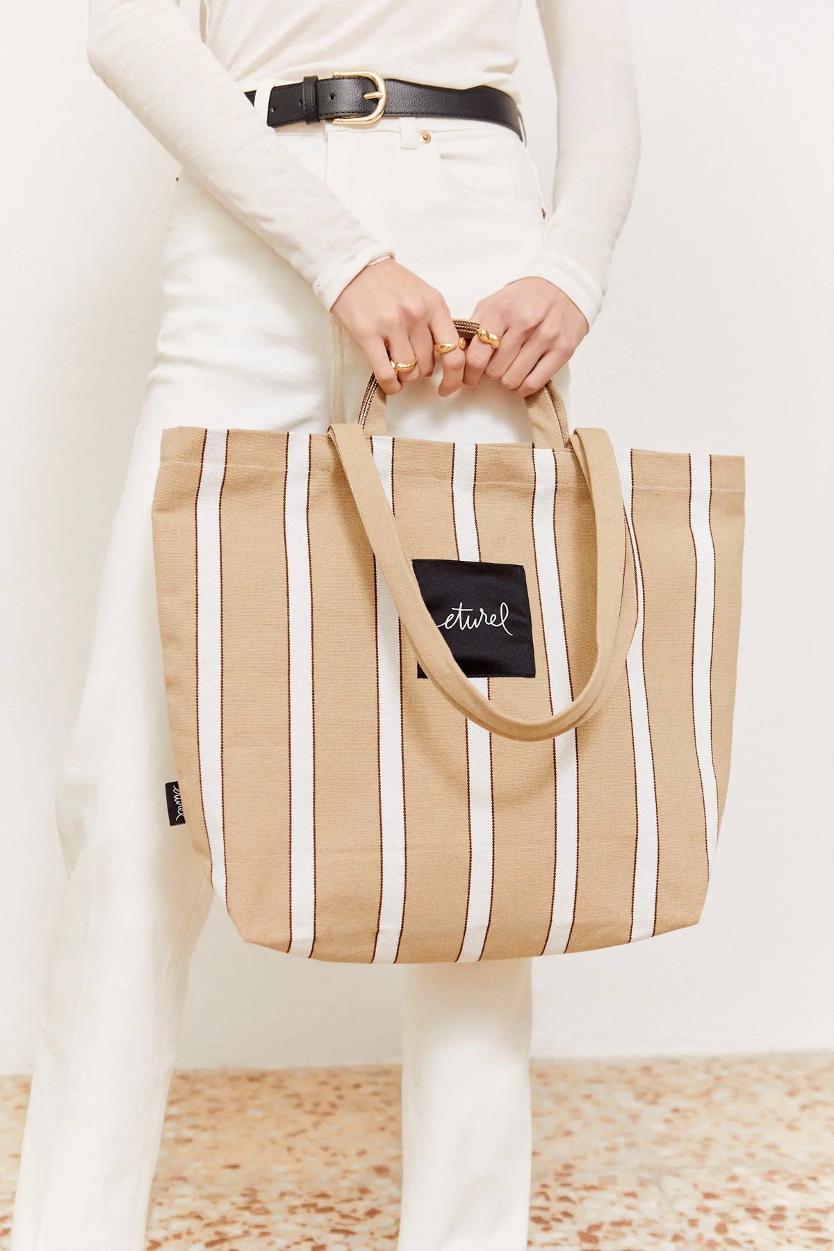 Beige and white striped canvas tote bag with dual shoulder straps and a black logo patch, held by a person.