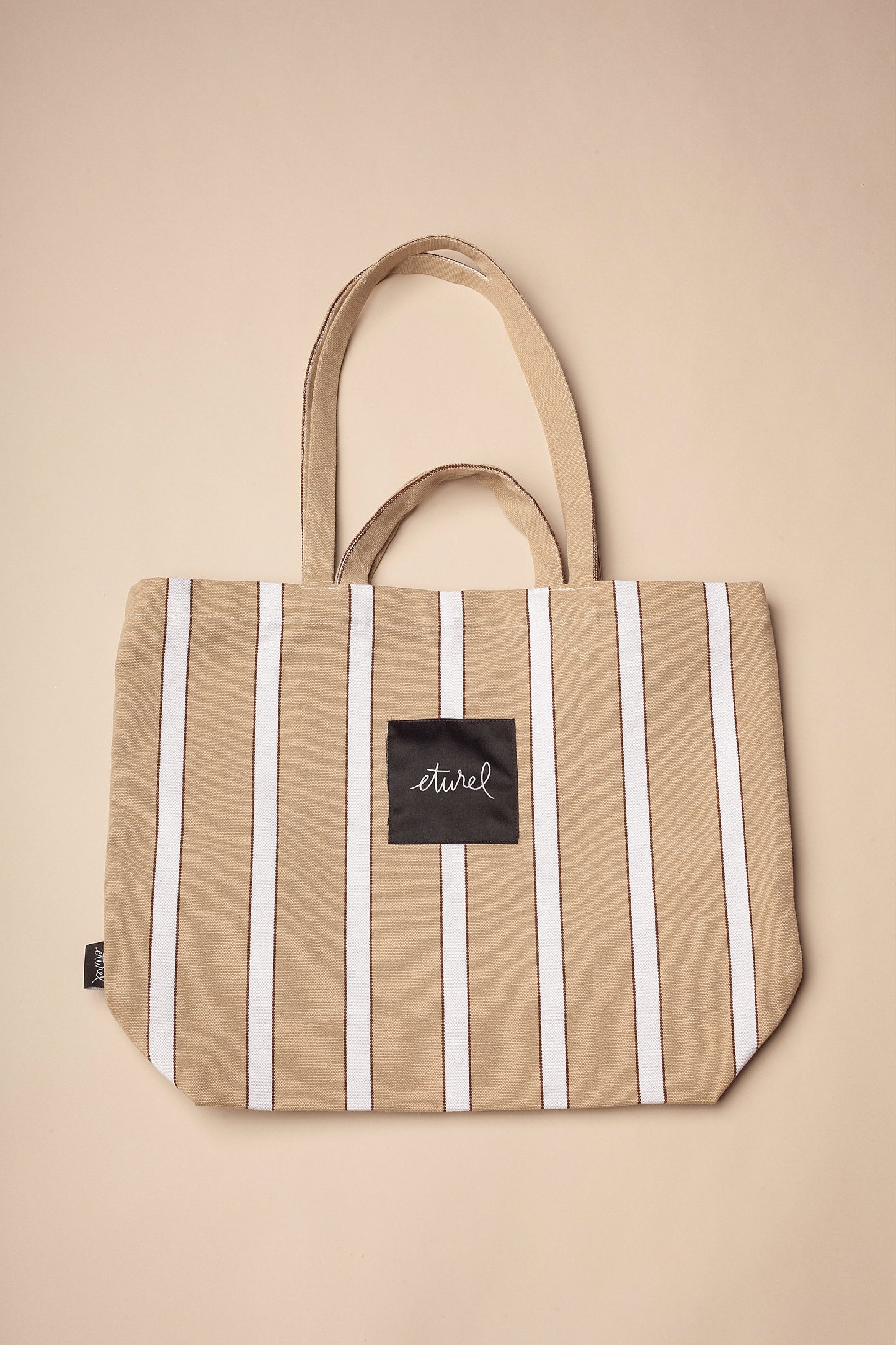 Bolso tote de lona estilo hamaca en beige con rayas verticales blancas, asas largas y etiqueta central rectangular.
