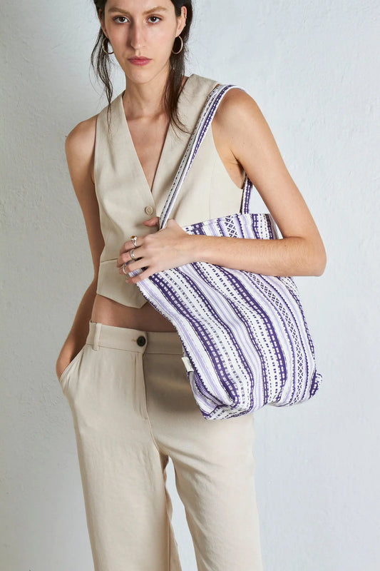 Tote bag de tela con estampado de rayas geométricas en tonos malva y blanco sobre modelo con chaleco y pantalón beige.